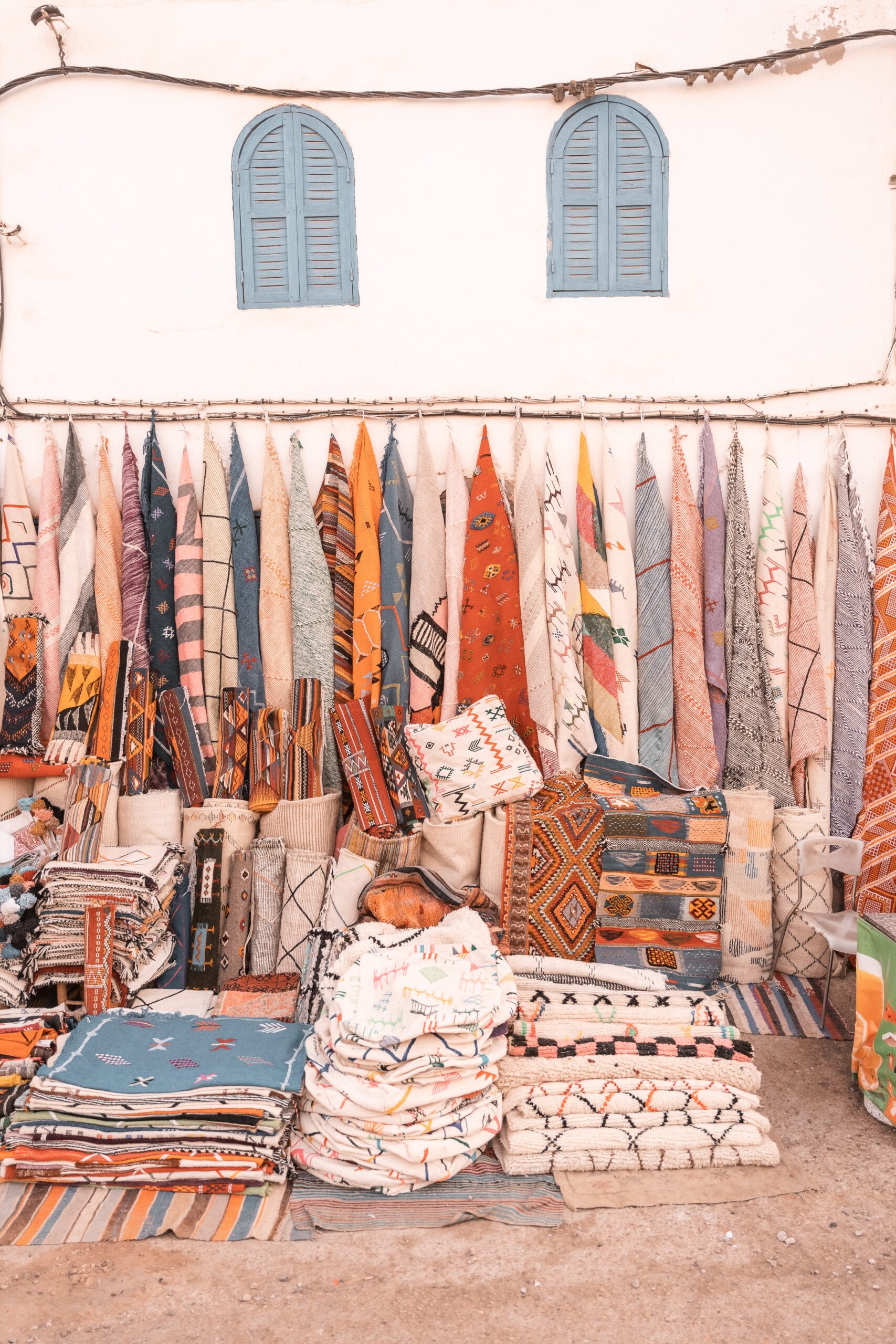 Handmade Moroccan carpets hanged on a white wall with two windows and a piles of handmade poufs and pillows put on the ground, reflecting the beauty of Moroccan handicraft