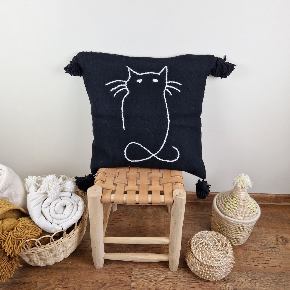 Handmade Black pillow with white cat drawing put on a handmade leather stool and a basket next to it full of handmade pompom blankets
