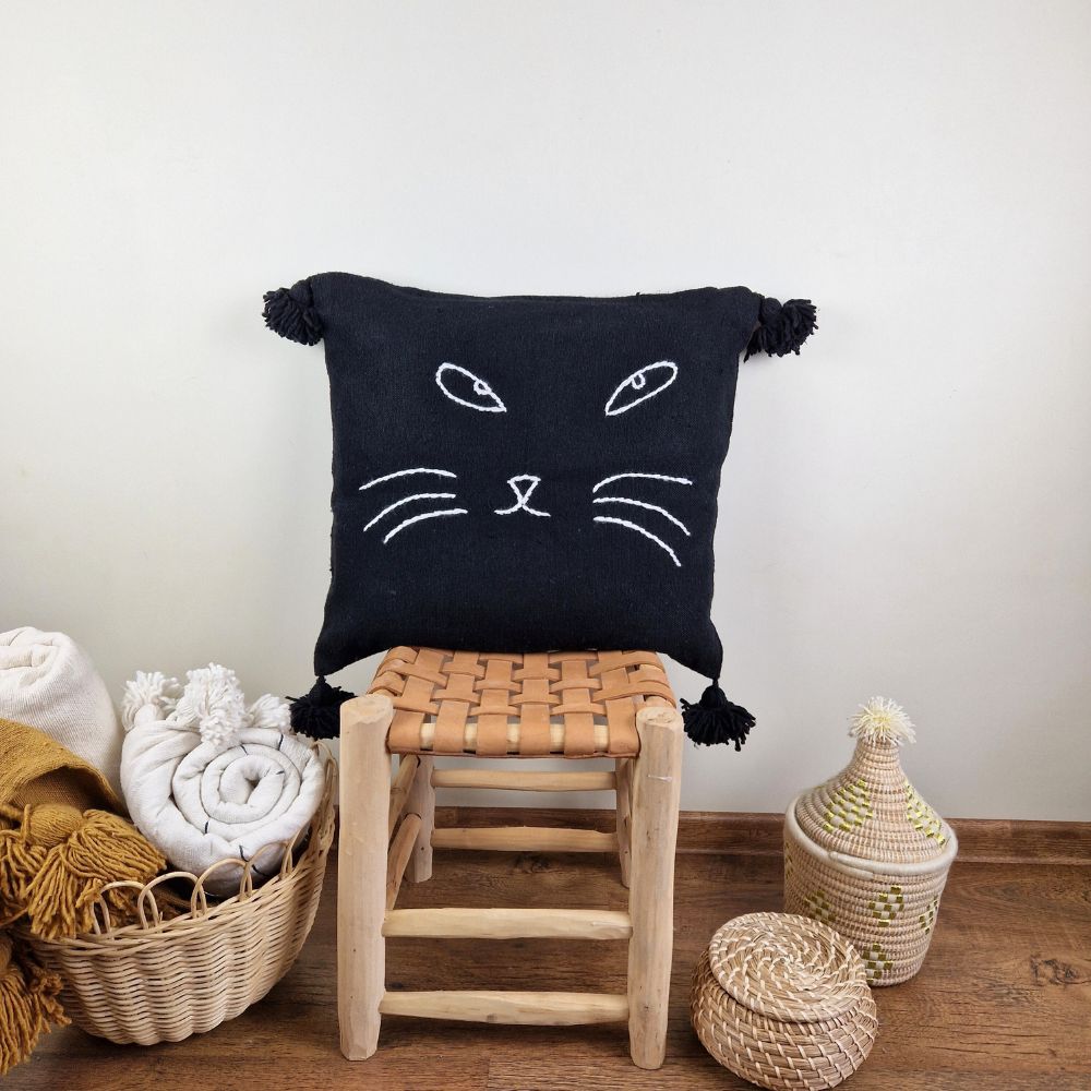 Handmade Black pillow with white cat face drawing put on a handmade leather stool and a basket next to it full of handmade pompom blankets