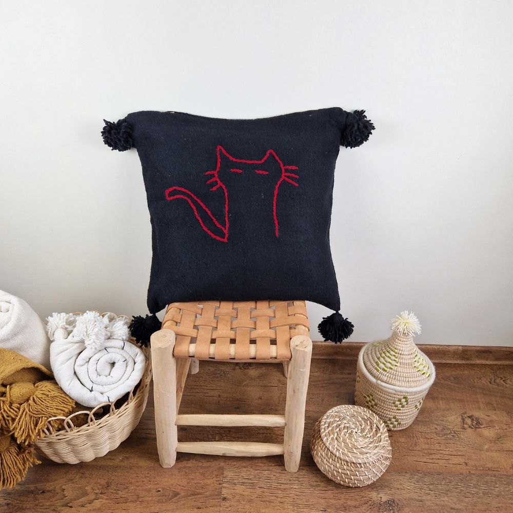 Handmade Black pillow with red cat drawing on a handmade leather stool and a basket next to it full of handmade pompom blankets