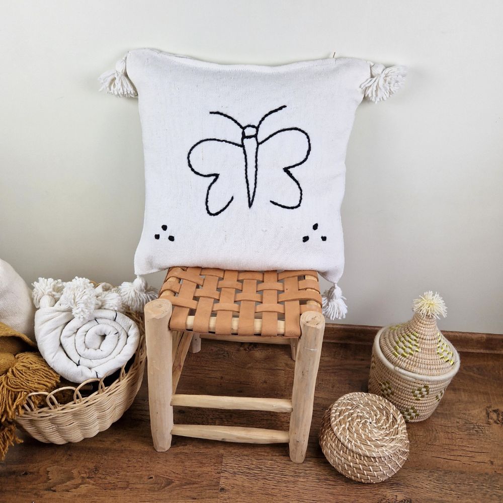 Handmade white pillow with black butterfly drawing on a handmade leather stool and a basket next to it full of handmade pompom blankets
