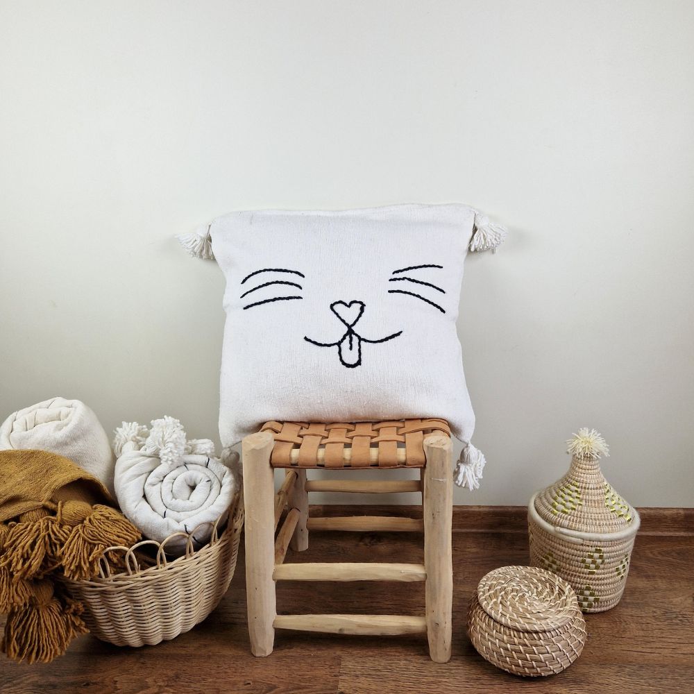 Handmade white pillow with cat face drawing on a handmade leather stool and a basket next to it full of handmade pompom blankets