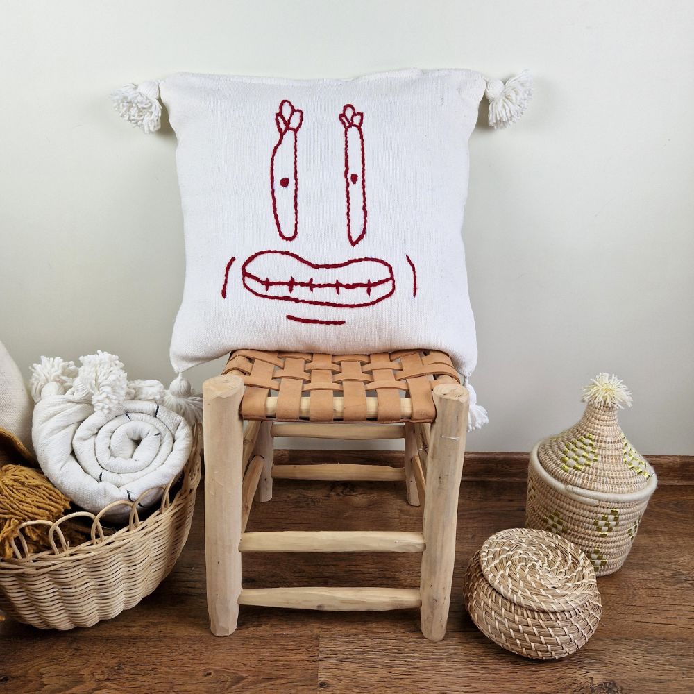 Handmade White pillow with Red Crab drawing on a handmade leather stool and a basket next to it full of handmade pompom blankets