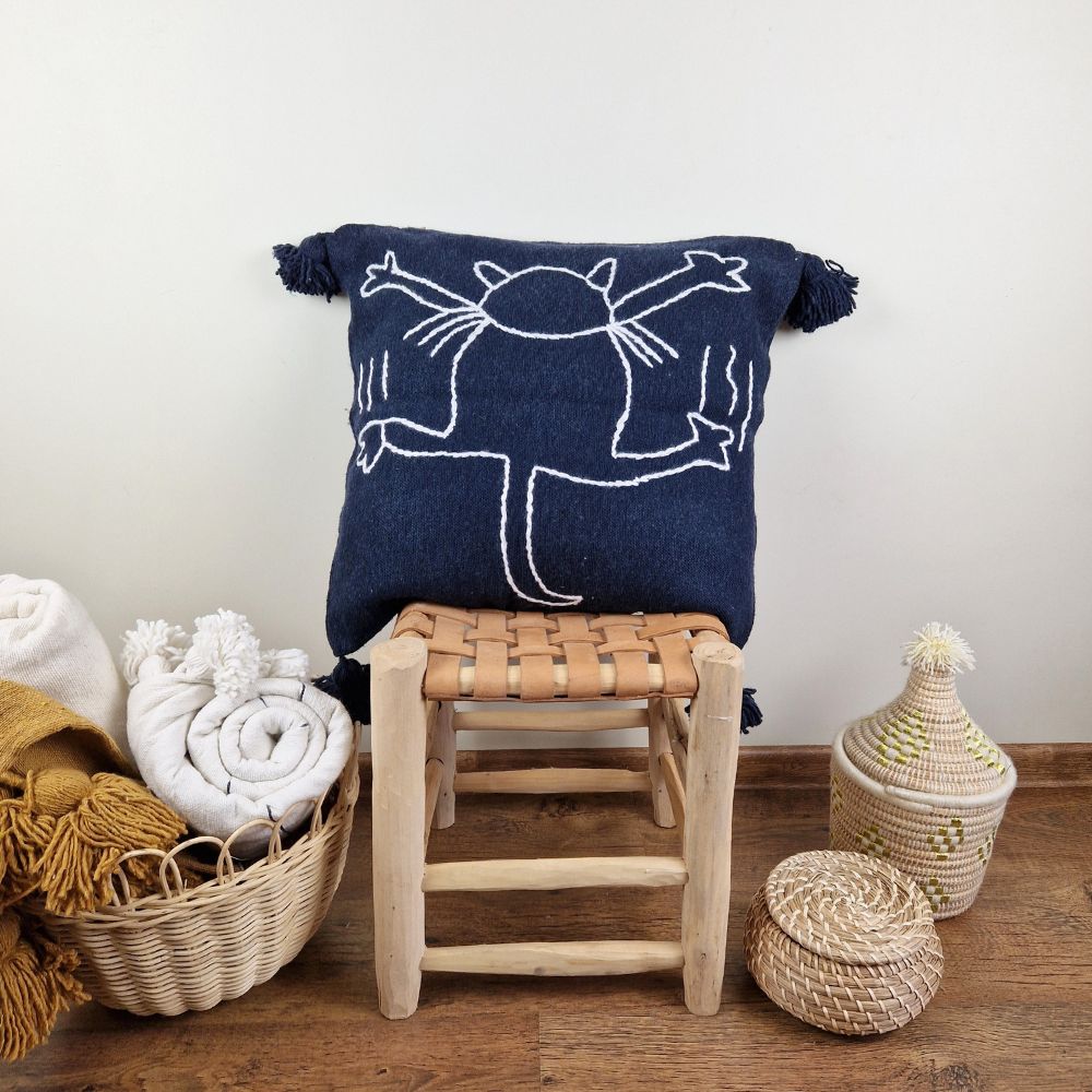 Handmade Dark Blue pillow with white cat drawing on a handmade leather stool and a basket next to it full of handmade pompom blankets