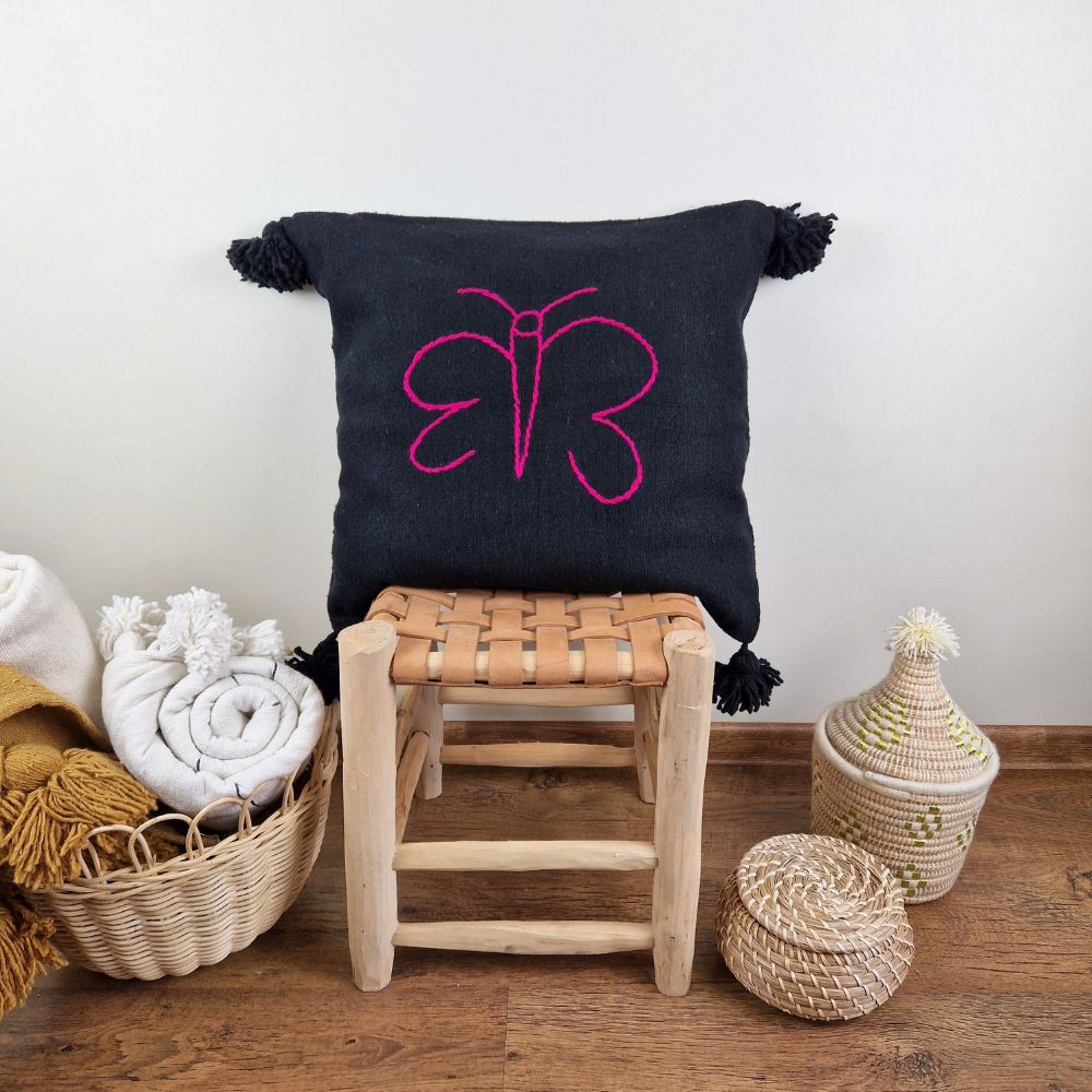 Handmade Black pillow with pink butterfly drawing on a handmade leather stool and a basket next to it full of handmade pompom blankets