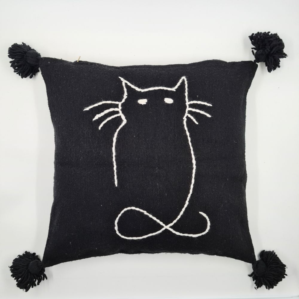 Handmade Black pillow with white cat drawing on a handmade leather stool and a basket next to it full of handmade pompom blankets