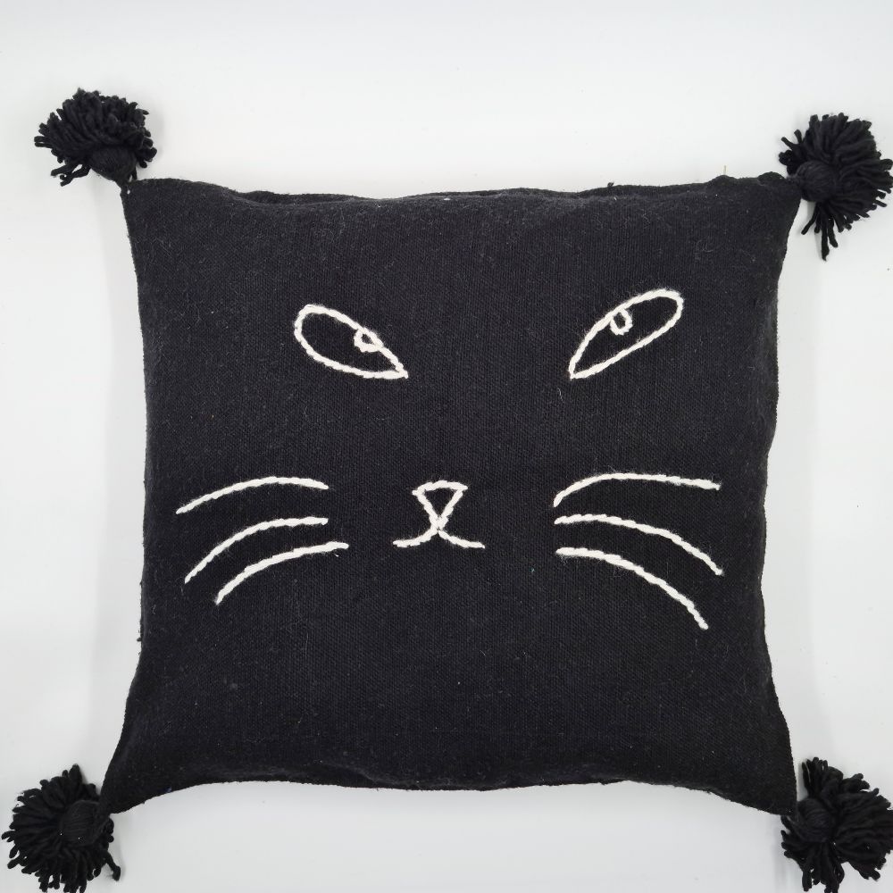 Handmade Black pillow with white cat face drawing put on a handmade leather stool and a basket next to it full of handmade pompom blankets