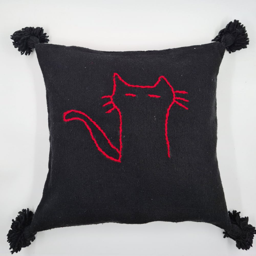 Handmade Black pillow with red cat drawing on a handmade leather stool and a basket next to it full of handmade pompom blankets