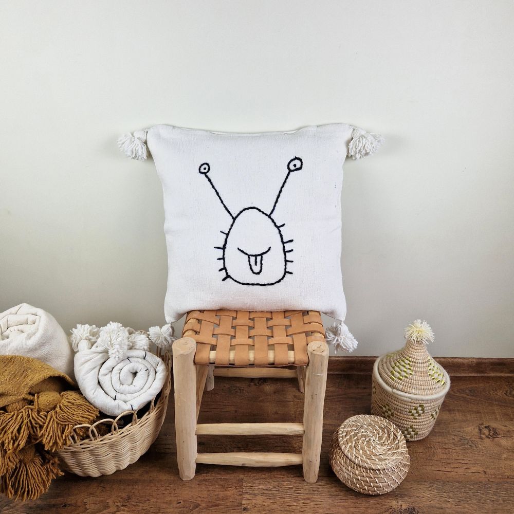 Handmade white pillow with black snail drawing on a handmade leather stool and a basket next to it full of handmade pompom blankets