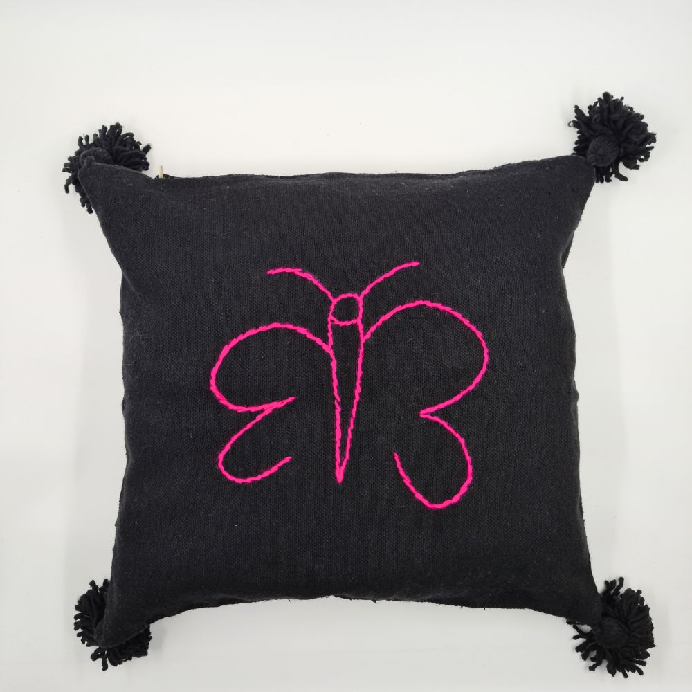 Black Handmade moroccan Pillow with pom poms and pink butterfly drawing put on a handmade leather stool and a basket next to it full of handmade pompom blankets